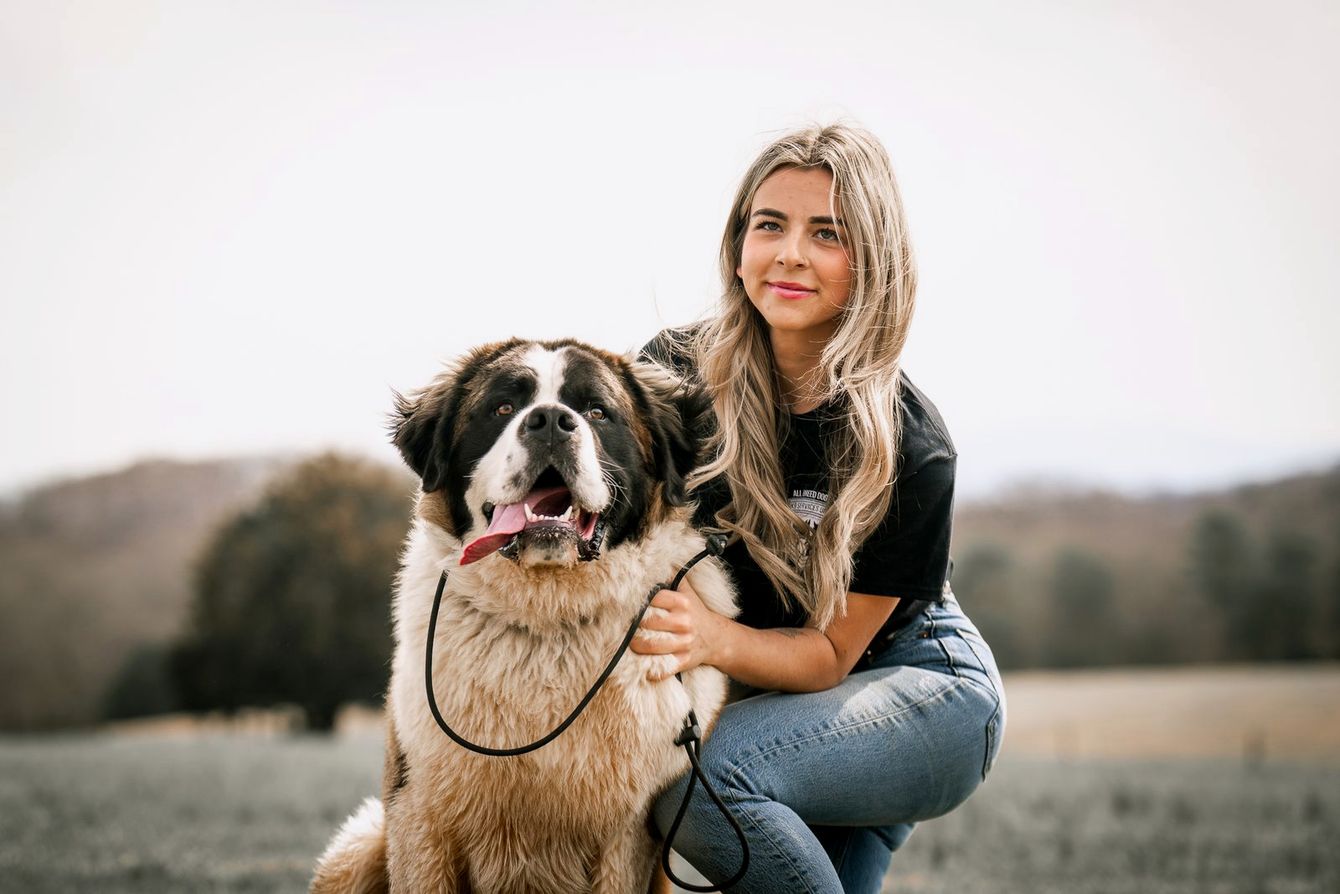 A trainer kneels with her arm around a smiling Saint Bernard in a hilltop field